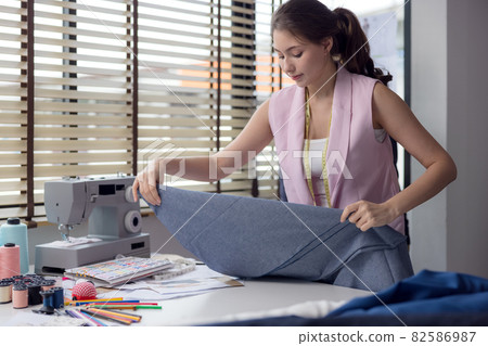 Portrait of a young, attractive woman on a stylish fashion designer's table, surrounded by color textile samples. Attractive woman working in a fashion studio with mannequins and colorful cloths. Portrait of a young, attractive woman on a stylish fashion designer's table, surrounded by color textile samples. Attractive woman working in a fashion studio with mannequins and colorful cloths. 82586987
