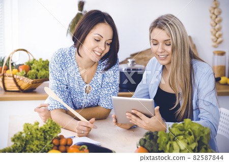 Two women friends choosing the recipe for a delicious meal while sitting at the table in the kitchen. Tablet pc is the best cookbook Two women friends choosing the recipe for a delicious meal while sitting at the table in the kitchen. Tablet pc is the best cookbook 82587844