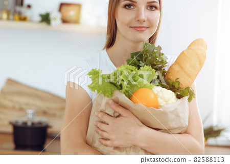 An attractive young woman holding the paper bag full of vegetables while standing and smiling in sunny kitchen. Cooking concept 82588113