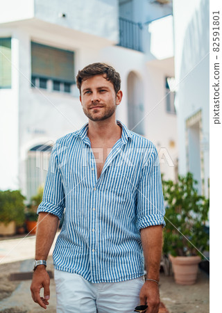 Adult male tourist in striped shirt looking away and dreaming while standing in sunlit yard during summer vacation Adult male tourist in striped shirt looking away and dreaming while standing in sunlit yard during summer vacation 82591801