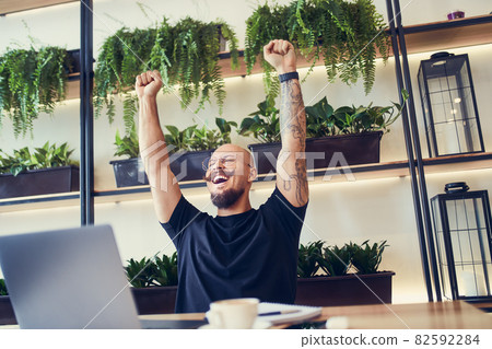 Happy young businessman sitting at laptop excited by good news online, lucky winner raising hand up Happy young businessman sitting at laptop excited by good news online, lucky winner raising hand up 82592284