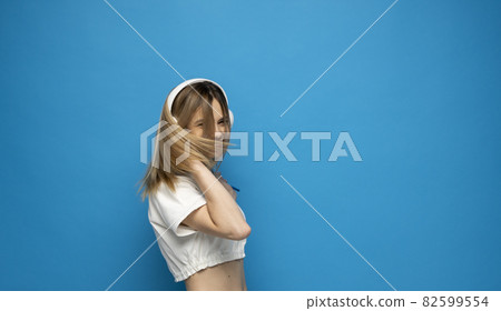 Beautiful attractive young blond woman wearing white t-shirt and glasses in white headphones listening music from smartphone, dancing and laughing on blue background in studio. Lifestyle. Beautiful attractive young blond woman wearing white t-shirt and glasses in white headphones listening music from smartphone, dancing and laughing on blue background in studio. Lifestyle. 82599554