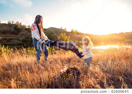 Global view, real life around the globe, local living. Mom and daughter in national clothes and flower wreaths are resting in meadow on sunset background Global view, real life around the globe, local living. Mom and daughter in national clothes and flower wreaths are resting in meadow on sunset background 82600645