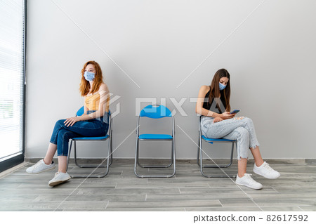 Two young woman with protection mask is sitting in a waiting hall with a distance. Corona virus outbreaking. 82617592