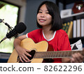 Happy cheerful pretty smiling of portrait a Asian girl vocalist with a Guitar recording a song front of microphone in living room. 82622296