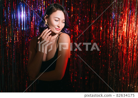 Merry christmas. Festive woman. Holiday look. Model shooting. Pretty smiling lady purple curly hair posing on shimmering red cascade curtain background blur shadow light copy space. 82634175