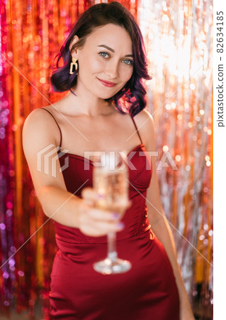 Christmas party. Festive woman. Holiday toast. Elegant happy lady purple curly hair cheering glass champagne on shimmering golden red cascade curtain decoration background blur light. 82634185