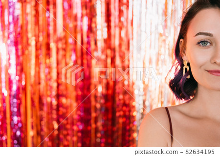 Christmas party. Festive woman. Stylish look. Pretty smiling lady purple curly hair posing on shimmering golden red cascade curtain decoration background copy space blur light. 82634195