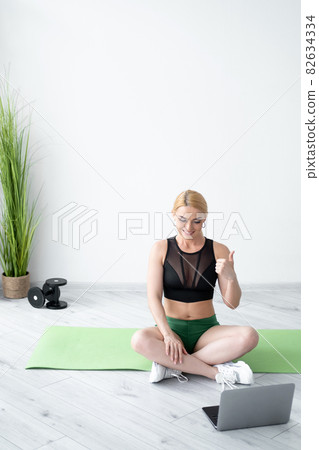Fitness joy. Supportive woman. Enjoying sport. Healthy body. Happy athletic lady in sportswear showing like gesture on laptop sitting yoga mat lotus pose in light room interior copy space. 82634334