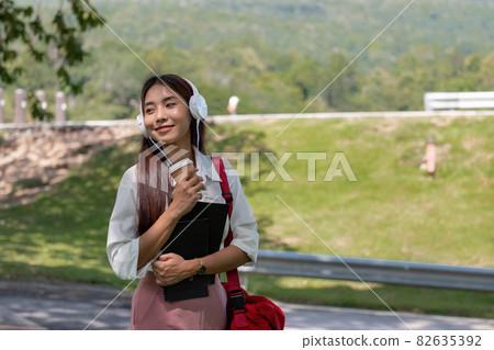 Happy young beautiful asian woman wearing headset and holding notebook in park. 82635392