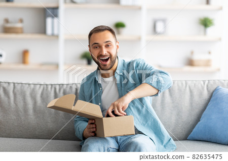 Portrait of cheerful young man receiving parcel, unboxing package, taking out gaming joystick, sitting on couch at home 82635475