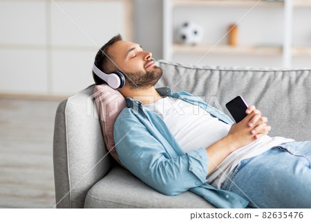 Relaxed young guy lying on couch in headphones, listening to music on smartphone, using new mobile app, mockup 82635476