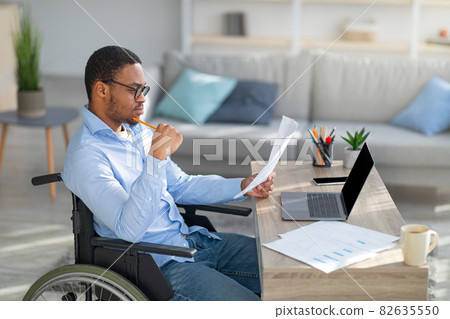 Focused black disabled man in wheelchair working with documents, using laptop at home office Focused black disabled man in wheelchair working with documents, using laptop at home office 82635550