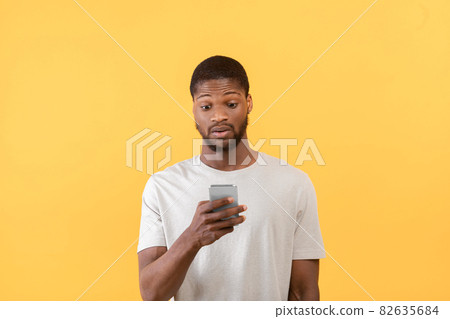 Emotional african american guy looking at smartphone, reading shocked message of promo or offer, yellow background Emotional african american guy looking at smartphone, reading shocked message of promo or offer, yellow background 82635684