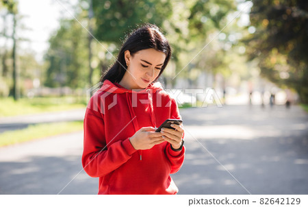 Stylish girl in red sweater posing on alley holding smartphone. Happy woman with beautiful smile chatting on cell smartphone while relaxing in the park, outdoors Stylish girl in red sweater posing on alley holding smartphone. Happy woman with beautiful smile chatting on cell smartphone while relaxing in the park, outdoors 82642129