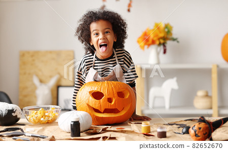 Playful little afro american boy holding carved orange pumpkin Jack o lantern while standing behind table 82652670