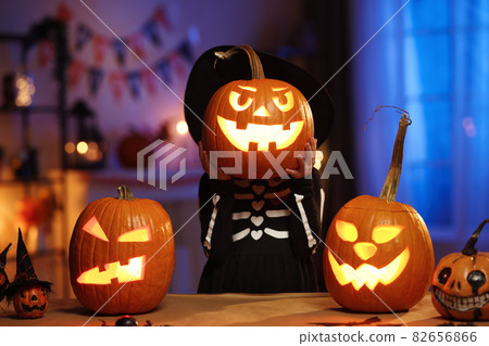 Little girl in Halloween costume holding glowing jack-o-lantern carved pumpkin in front of face 82656866