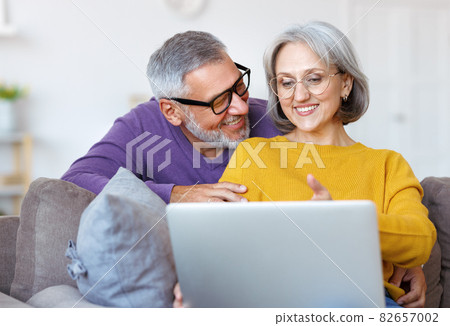 Beautiful mature family couple looking at laptop with smile on face while spending time together Beautiful mature family couple looking at laptop with smile on face while spending time together 82657002