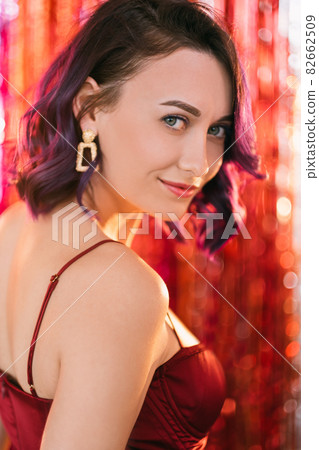 Christmas look. Glamorous woman. Party celebration. Elegant happy lady purple curly hair posing on shimmering golden red cascade curtain decoration background blur light. 82662509