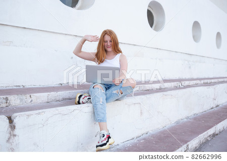 Red-haired teen girl talking on video call while sitting on steps with laptop Red-haired teen girl talking on video call while sitting on steps with laptop 82662996