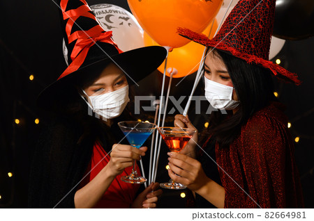 Two beautiful young woman in witch halloween costumes drinking cocktail and holding balloons wearing protection face mask against coronavirus (covid-19) in halloween party. Two beautiful young woman in witch halloween costumes drinking cocktail and holding balloons wearing protection face mask against coronavirus (covid-19) in halloween party. 82664981