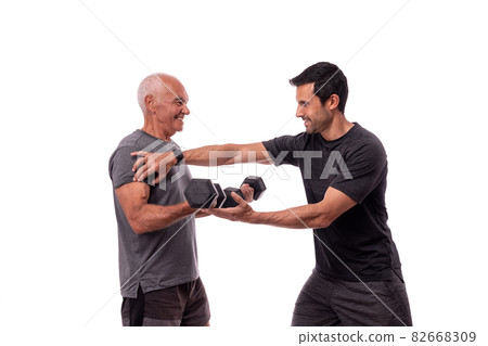 An elderly European man, with a personal trainer, trains, lifting weights, on a white background. 82668309
