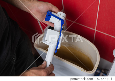 Toilet tank parts replacement. A man in orange gloves repairs the toilet tank drain. 82668401