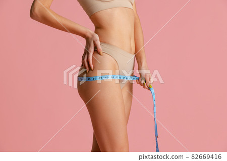 Close-up slim female body with well kept skin in pastel color panties using centimeter isolated over pink studio background. 82669416