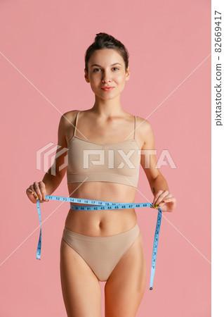 Portrait of young beautiful slim girl in lingerie takes measurements from yourself isolated over pink studio background. 82669417