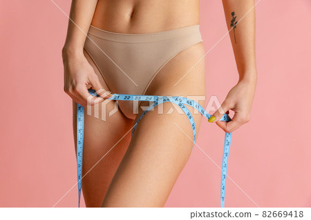 Close-up slim female body with well kept skin in pastel color panties using centimeter isolated over pink studio background. 82669418