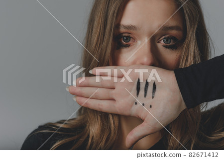 A crying young girl covering her mouth with her palm, on which there are three exclamation marks. 82671412