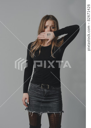A crying young girl covering her mouth with her palm, on which there are three exclamation marks. 82671426
