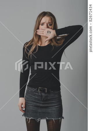 A crying young girl covering her mouth with her palm, on which there are three exclamation marks. 82671434