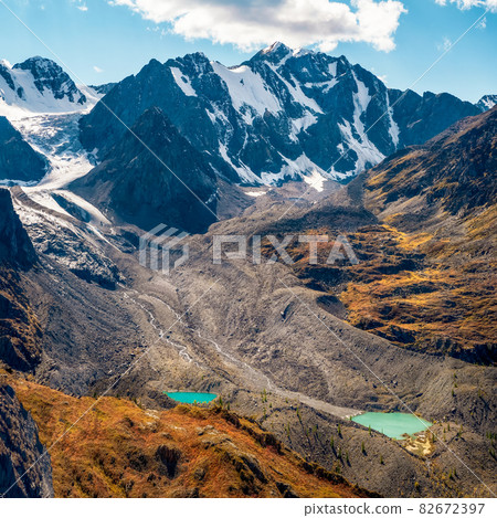 Awesome alpine panoramic view from pass to mountain valley in sunlight and great mountain silhouettes on horizon. Colorful green landscape with silhouettes of big rocky mountains and epic deep gorge. Awesome alpine panoramic view from pass to mountain valley in sunlight and great mountain silhouettes on horizon. Colorful green landscape with silhouettes of big rocky mountains and epic deep gorge. 82672397