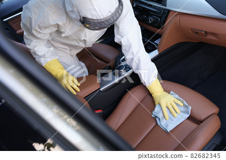 Man in protective suit and rubber gloves wiping leather car seat with rag with disinfectant closeup 82682345