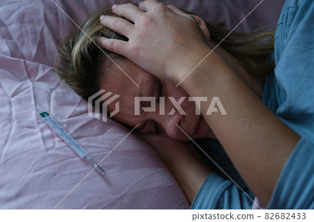 Woman lying in bed and holding sore head with hands near mercury thermometer 82682433