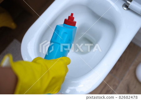 Cleaner in yellow rubber gloves pouring detergent under rim of toilet closeup 82682478