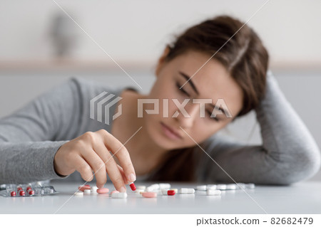 Indifferent caucasian young female suffering from depression, touching many scattered pills on table 82682479