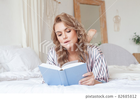 White cozy bed and a beautiful woman reading a book, concepts of home and comfort White cozy bed and a beautiful woman reading a book, concepts of home and comfort 82685812