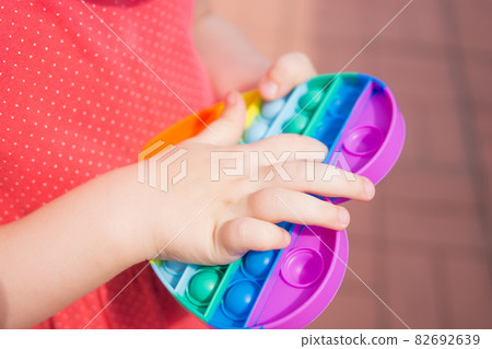 Children's hands hold a silicone toy. Kid having fun with popular simple dimple multicolored rainbow colors toy. Kids being kids LGBTQ colors Vertical. Children's hands hold a silicone toy. Kid having fun with popular simple dimple multicolored rainbow colors toy. Kids being kids LGBTQ colors Vertical. 82692639