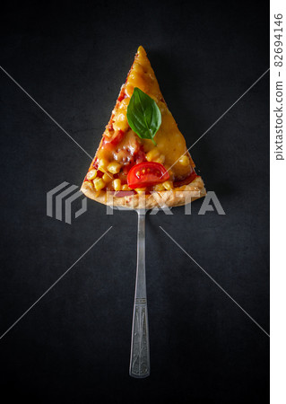Slice of pizza with tomatoes and basil on a black background. National Italian food, pizza with tomatoes, corn, green basil and cheese on a round board. Vertical photo, place for text. 82694146
