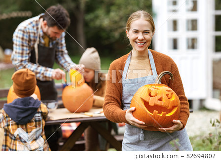 Smiling woman with jack-o-lantern smiling at camera while carving pumpkins with family in backyard Smiling woman with jack-o-lantern smiling at camera while carving pumpkins with family in backyard 82694802