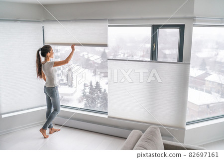 Woman opening home curtains in urban condo. Modern top down bottom up privacy cellular shades on apartment window keeping heat in winter with honeycomb blind curtain. Cordless pleated shades 82697161