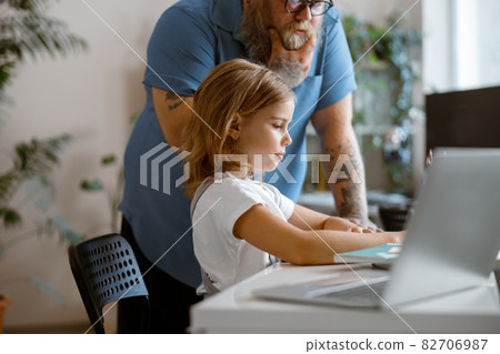 Positive little girl does homework while thoughtful daddy stands nearby in light room Positive little girl does homework while thoughtful daddy stands nearby in light room 82706987