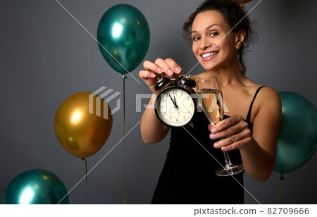Beautiful woman in evening black dress holds a flute of sparkling wine and shows an alarm clock isolated over gray background with inflated air balloons. Christmas and New Year concept, Anniversary. 82709666