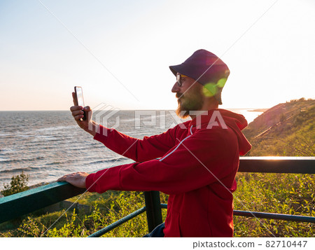 Bearded millennial man in red hoody backpack on bench with smartphone taking photo of autumn sea view. Bearded millennial man in red hoody backpack on bench with smartphone taking photo of autumn sea view. 82710447