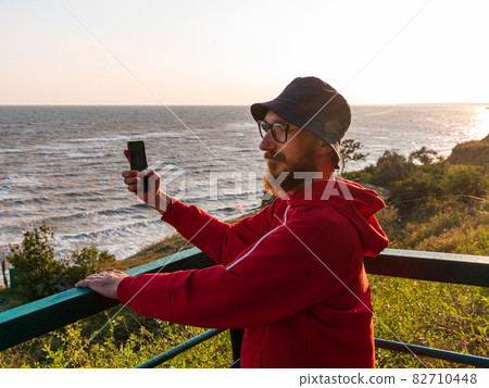 Bearded millennial man in red hoody backpack on bench with smartphone taking photo of autumn sea view. Bearded millennial man in red hoody backpack on bench with smartphone taking photo of autumn sea view. 82710448