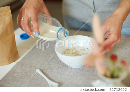 Female hand holding bottle pouring milk in cereal granola flakes bowl Female hand holding bottle pouring milk in cereal granola flakes bowl 82712022