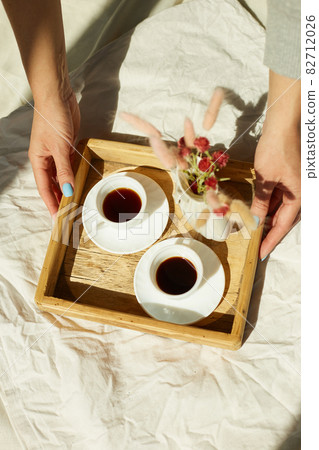 Breakfast in bed, female hands put try with two cups coffee and flower in sunlight Breakfast in bed, female hands put try with two cups coffee and flower in sunlight 82712026