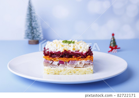 Herring salad under a fur coat. Traditional Russian multilayered salad from herring, beets, potatoes, carrots and eggs. Close-up, blue background., miniature Christmas tree. 82722171